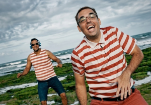 O humorista Tenóbio (Gabriel Bandarra) vestindo camisa listrada em uma praia; imagem ilustra a paródia satírica 'Em cima do triplex' sobre o Governo Lula.