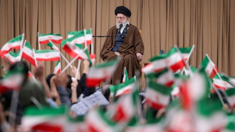 Líder Supremo Ali Khamenei em um púlpito com microfones, cercado por bandeiras do Irã; imagem ilustra o anúncio de sucessão em Teerã.