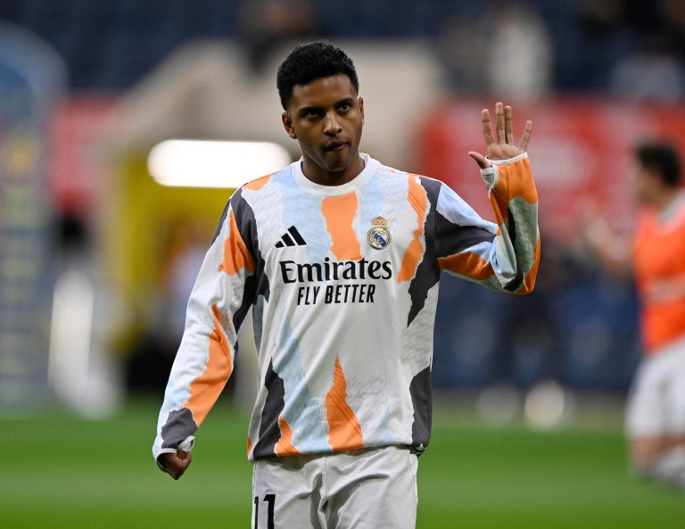 Atacante Rodrygo Goes com o uniforme do Real Madrid, fazendo um gesto com a mão durante partida; a imagem ilustra a notícia sobre sua cirurgia de joelho e ausência na Copa do Mundo de 2026.