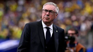 O técnico Carlo Ancelotti, vestindo terno preto, camisa branca e gravata escura com o broche da CBF na lapela, olha para o horizonte com expressão séria em um estádio lotado com torcedores ao fundo.