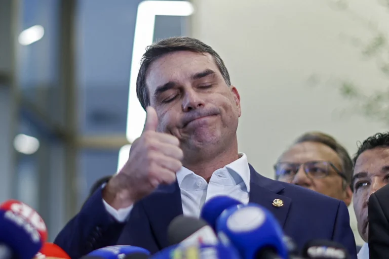 O senador Flávio Bolsonaro, vestindo paletó azul e camisa branca, gesticula com a mão direita erguida enquanto fala ao microfone durante pronunciamento oficial.