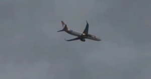 Avião comercial bimotor de fuselagem branca e cauda alaranjada voando contra o céu nublado com chamas visíveis sob o motor direito após colisão com pássaro.