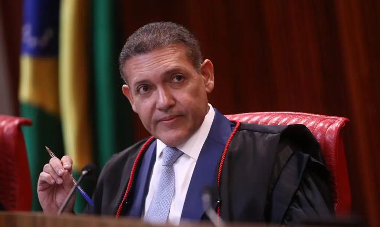 O ministro Kassio Nunes Marques vestindo toga preta, sentado em uma cadeira vermelha no plenário com a bandeira do Brasil desfocada ao fundo.