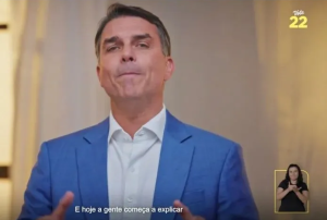 O candidato Flávio Bolsonaro vestindo terno azul e camisa branca gesticulando durante programa eleitoral, com a intérprete de libras no canto inferior direito, o número 22 no canto superior direito e a legenda 