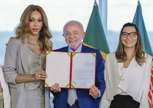 Erika Hilton à esquerda com terno cinza segurando um documento de capa vermelha ao lado de Luiz Inácio Lula da Silva ao centro de terno azul e Janja da Silva à direita sorridente com casaco branco.