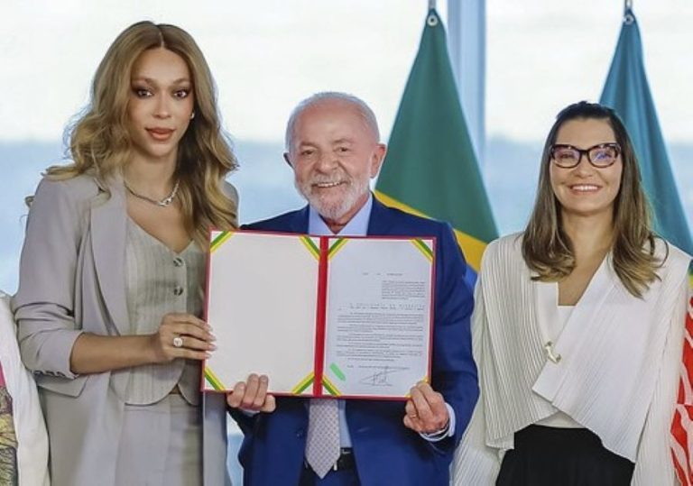 Erika Hilton à esquerda com terno cinza segurando um documento de capa vermelha ao lado de Luiz Inácio Lula da Silva ao centro de terno azul e Janja da Silva à direita sorridente com casaco branco.