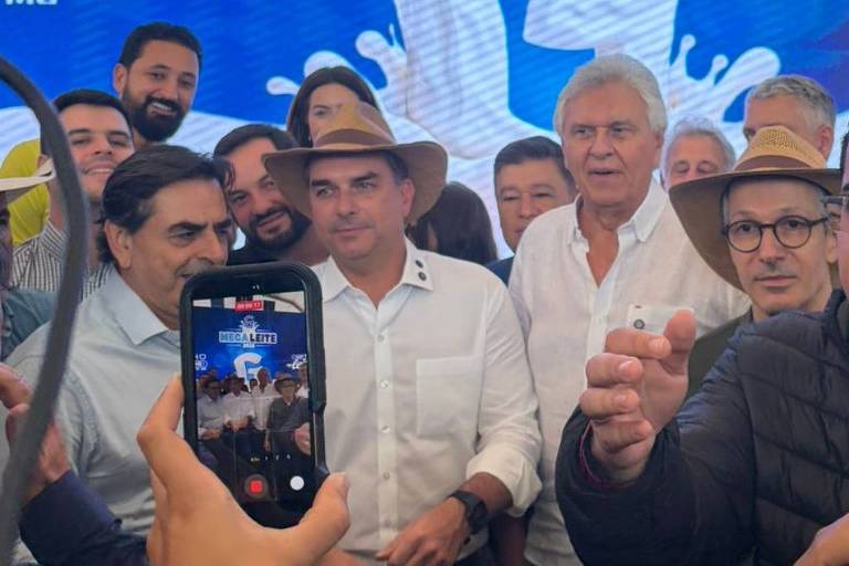 Romeu Zema à esquerda usando óculos e chapéu claro de aba, Flávio Bolsonaro ao centro de camisa branca e Ronaldo Caiado à direita sorrindo em evento político.