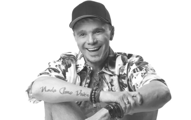 Fotografia em preto e branco do cantor Netinho sorrindo, vestindo um boné escuro e camisa estampada, com os braços cruzados apoiados sobre as pernas e a tatuagem