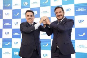 Ciro Nogueira à esquerda e Antônio Rueda à direita, ambos de terno azul e sorridentes, apertam as mãos em frente a painel com logotipos da federação União Progressista.